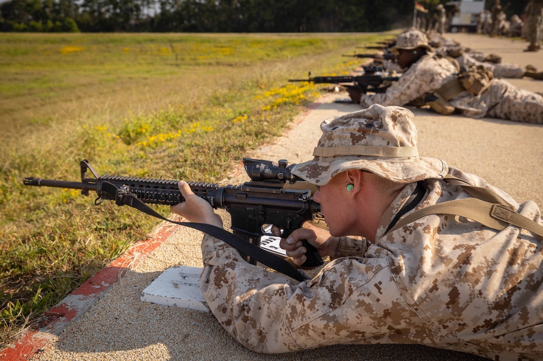 Marines lie on the ground while firing weapons into a field during the day.