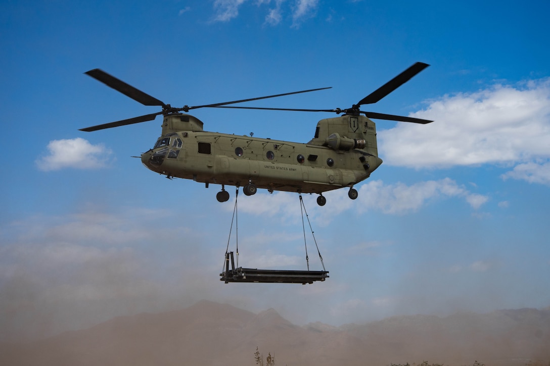 A helicopter with a sling-loaded platform hanging from it flies against a blue sky over dust clouds, with mountains in the distance.