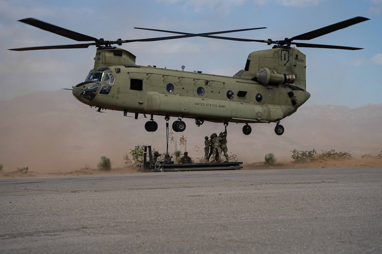 A group of soldiers holds on to a sling load while standing on a platform under a helicopter in flight during the day, with haze in the background.