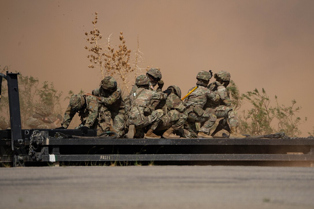 A group of helmeted soldiers in camouflage stand on a metal platform with vegetation and dust clouds behind.