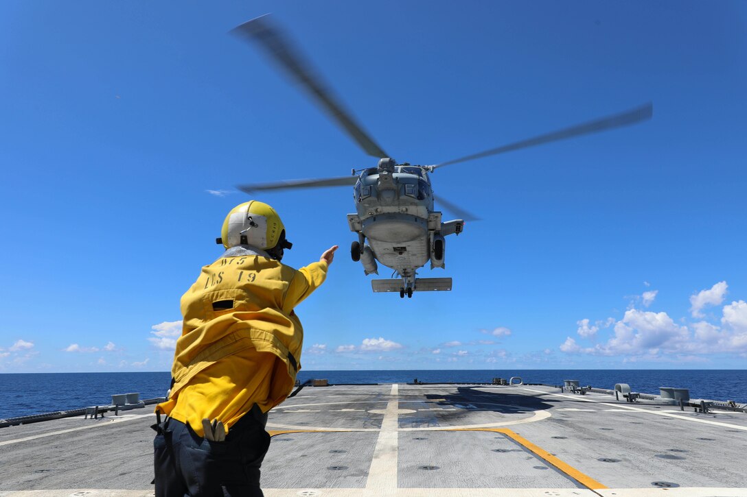 A sailor in a yellow jacket and helmet signals toward a helicopter hovering over a ship deck with the ocean behind, under blue skies.