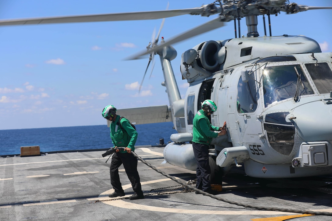 Two people in green jackets and helmets stand on a ship deck during the day, while one holds a hose and the other touches a parked helicopter.