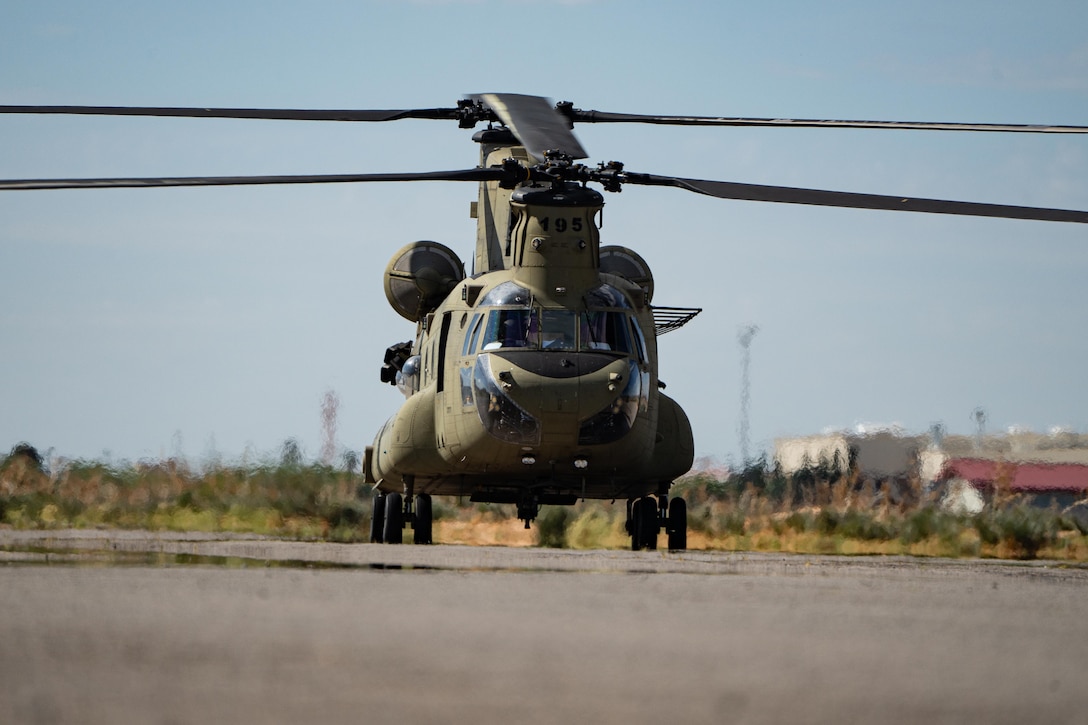 An olive-colored helicopter is parked on a runway during the day with some vegetation and a building behind.