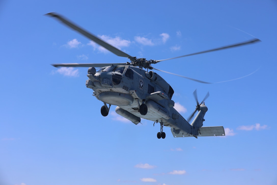 A gray helicopter flies against a blue sky during the day with a few clouds visible behind.
