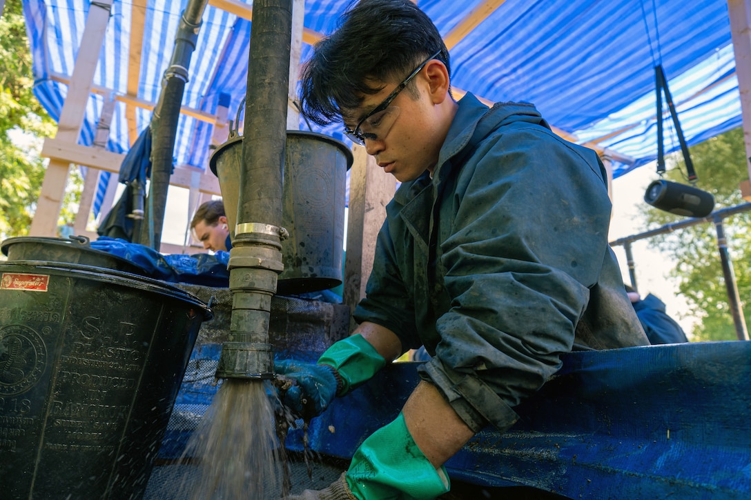 An airman holds the nozzle of a large hose attached to a pipe above and spews water to screen debris in a basin, with buckets and other people nearby on a sunny day outdoors.