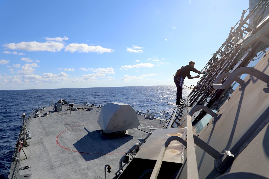 A sailor balances on metal rungs above a ship deck while cleaning windows under a blue sky, with the ocean behind.