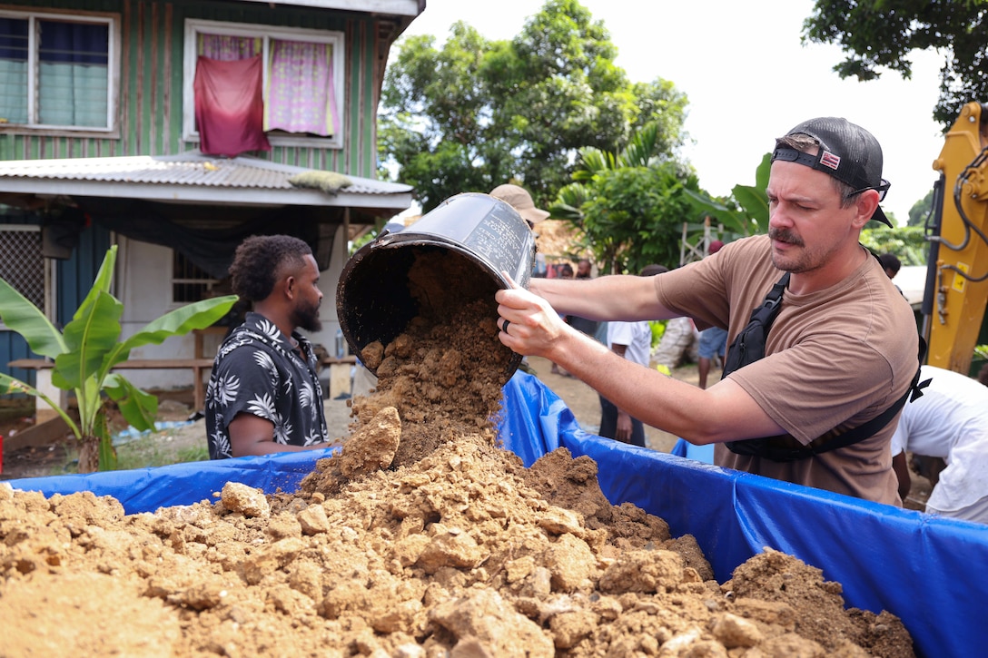U.S. service members and other people use buckets to collect soil in a large bin lined with blue material, with a house and trees in the background during daytime.