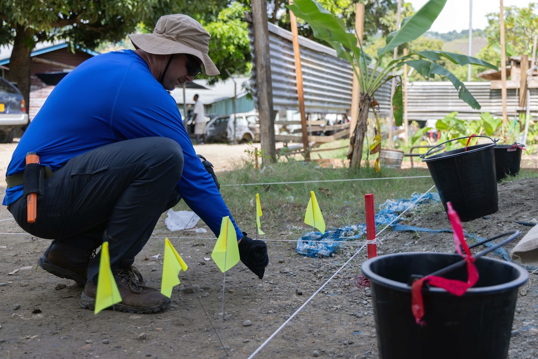 A Marine squats to place small yellow flags in a dirt area marked with string, with houses, a person, green vegetation and a metal gate in the background on a sunny day.
