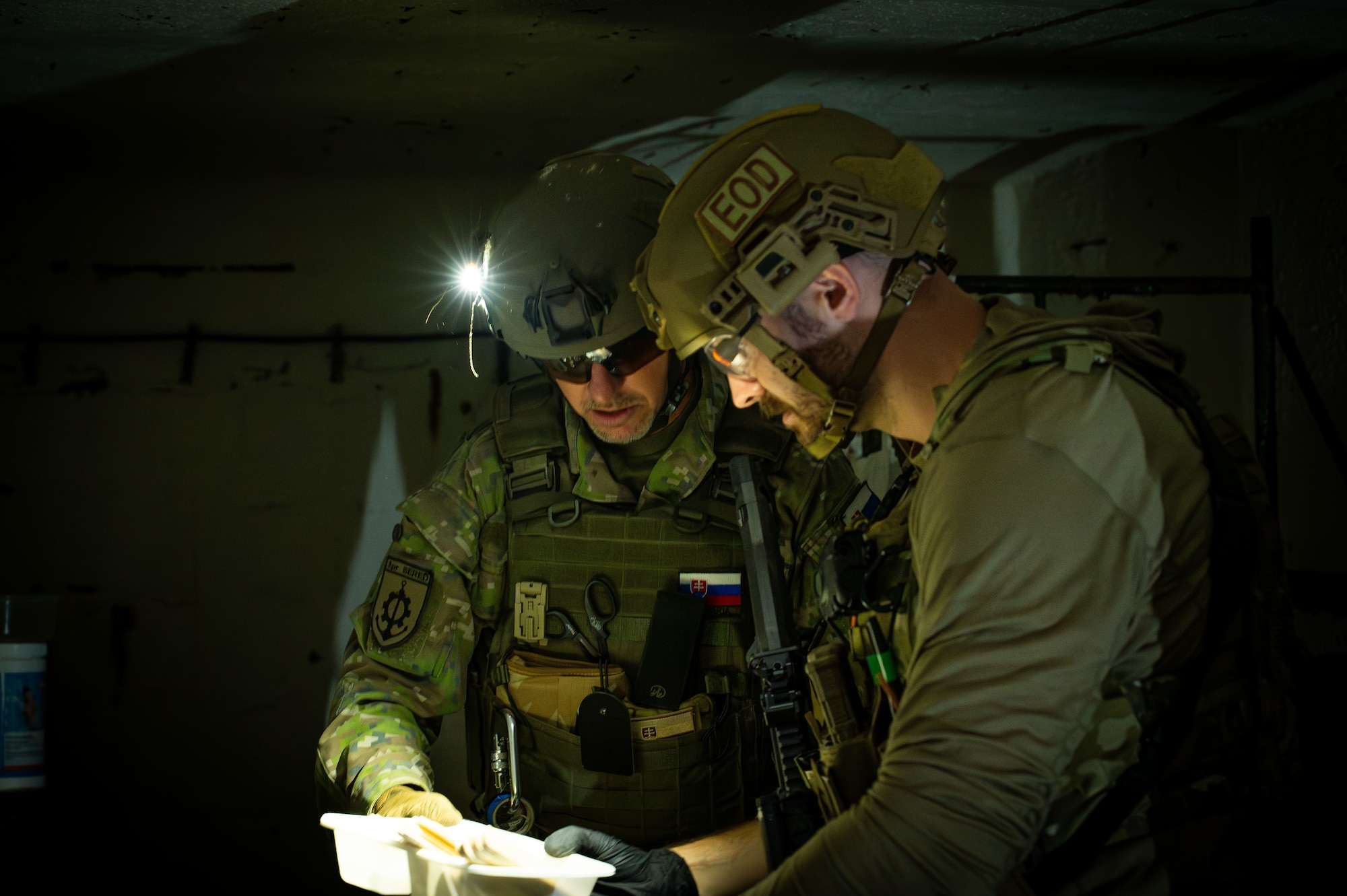 An Airman and a member of the Slovak Armed Forces in a dark room with flashlights looking at a plastic bag.