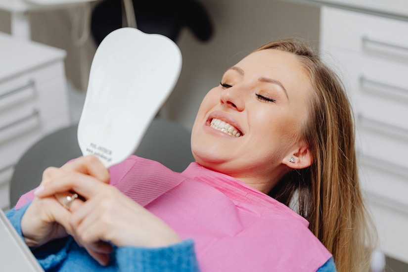 Woman in a dentist's chair holds a mirror and looks at her teeth.