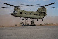 A group of soldiers holds on to a sling load while standing on a platform under a helicopter in flight during the day, with haze in the background.
