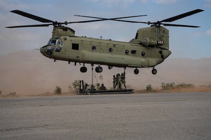 A group of soldiers holds on to a sling load while standing on a platform under a helicopter in flight during the day, with haze in the background.
