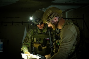 An Airman and a member of the Slovak Armed Forces in a dark room with flashlights looking at a plastic bag.
