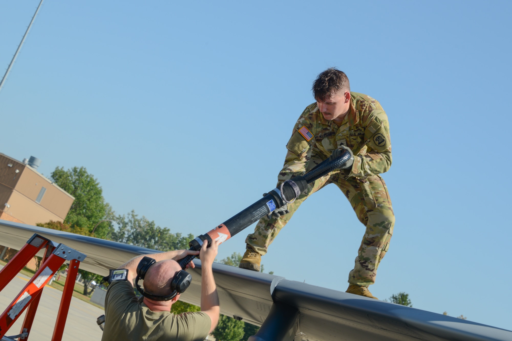 A Soldier standing atop the wing of a U.S. Air Force A-7D Corsair II fighter jet receives sling load gear from a service member on the ground.