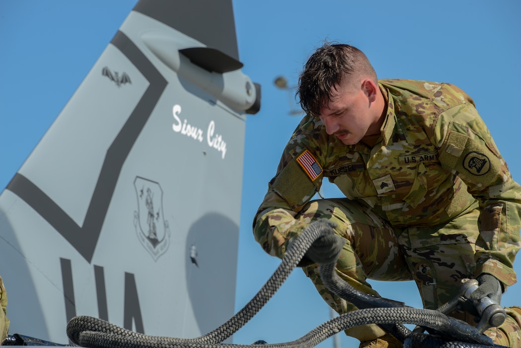 Soldier of Bravo Company, 1st Battalion, 171st Aviation Regiment, kneels atop the wings of a U.S. Air Force A-7D Corsair II fighter jet to rig it with sling load gear.