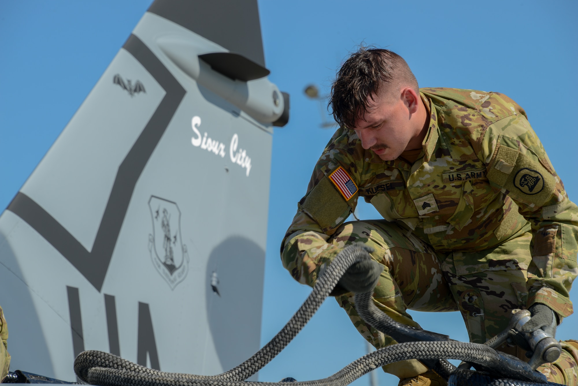 Soldier of Bravo Company, 1st Battalion, 171st Aviation Regiment, kneels atop the wings of a U.S. Air Force A-7D Corsair II fighter jet to rig it with sling load gear.