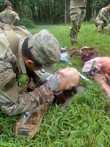 A Soldier simulates a medical procedure on a mannequin in a grassy field.