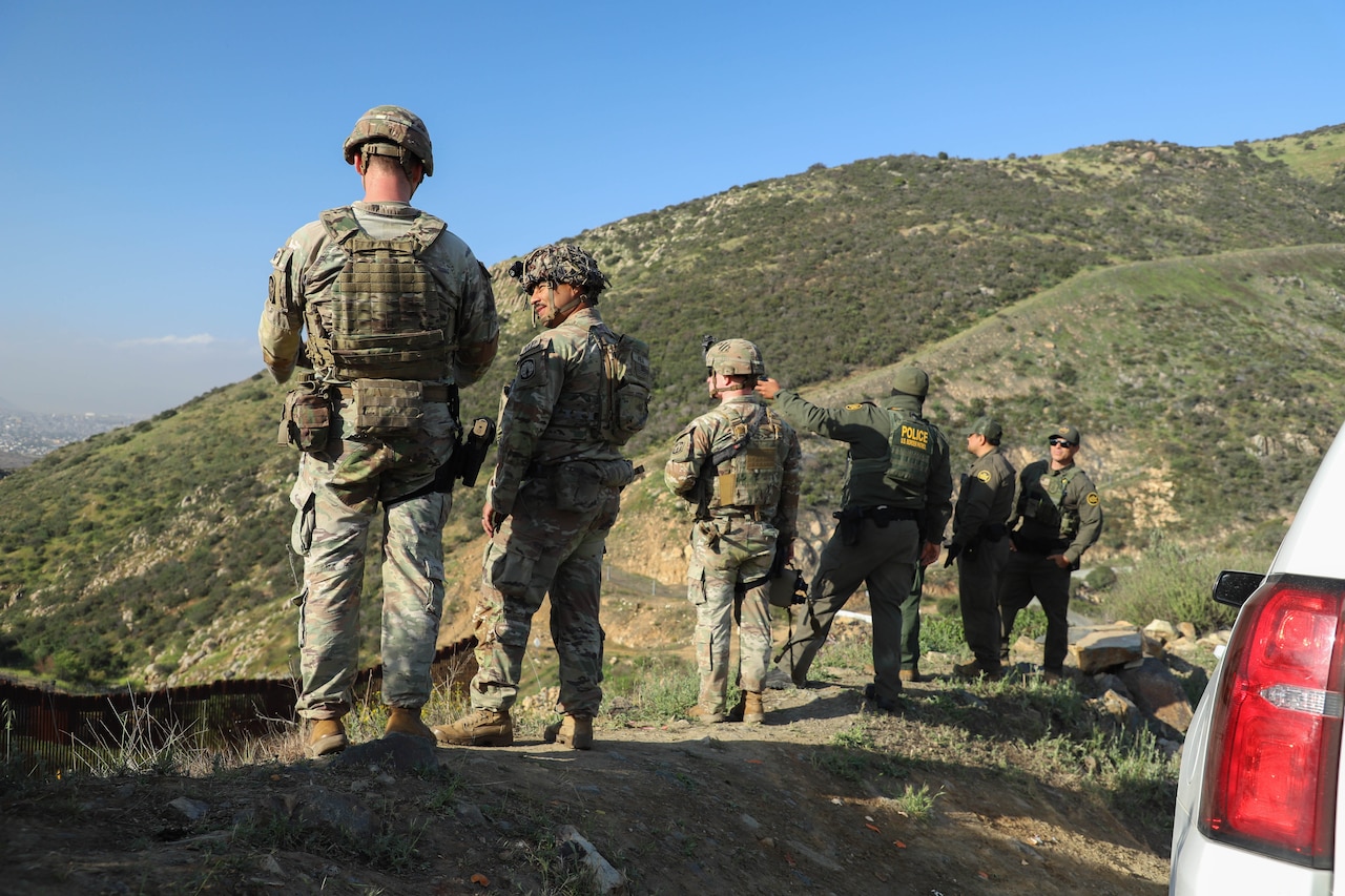 Three soldiers wearing camouflage military uniforms stand on a hill next to three other people in green police uniforms. One of the men is pointing toward the valley below.