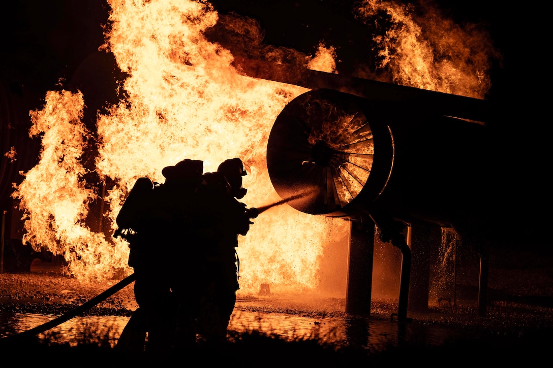 Firefighters in silhouette use a hose to put out an aircraft fire in the dark.