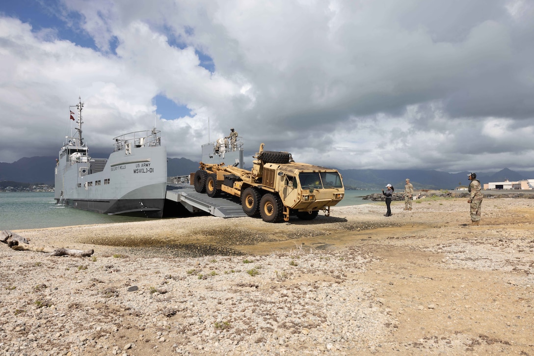 Soldiers on a boat offload a military vehicle onto a beach under a cloudy sky with mountains in the distance.