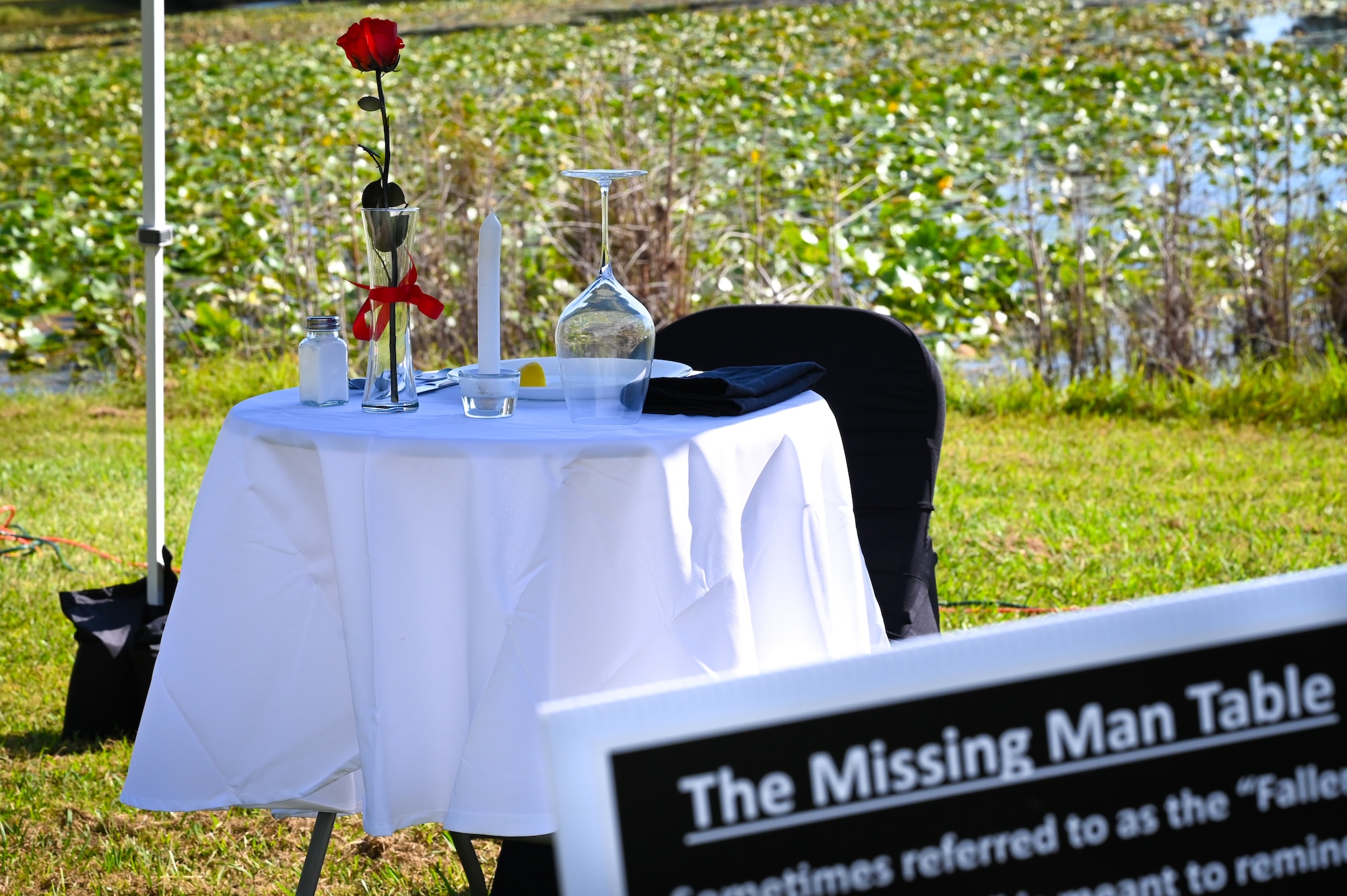 A missing man table composed of a small table set for one with a white tablecloth, a single rose in a vase, a yellow ribbon, a slice of lemon on a small bread plate, a salt shaker, an inverted glass, a lit candle and an empty chair. A poster is in the foreground with the title "The Missing Man Table." There is a pond in the background.