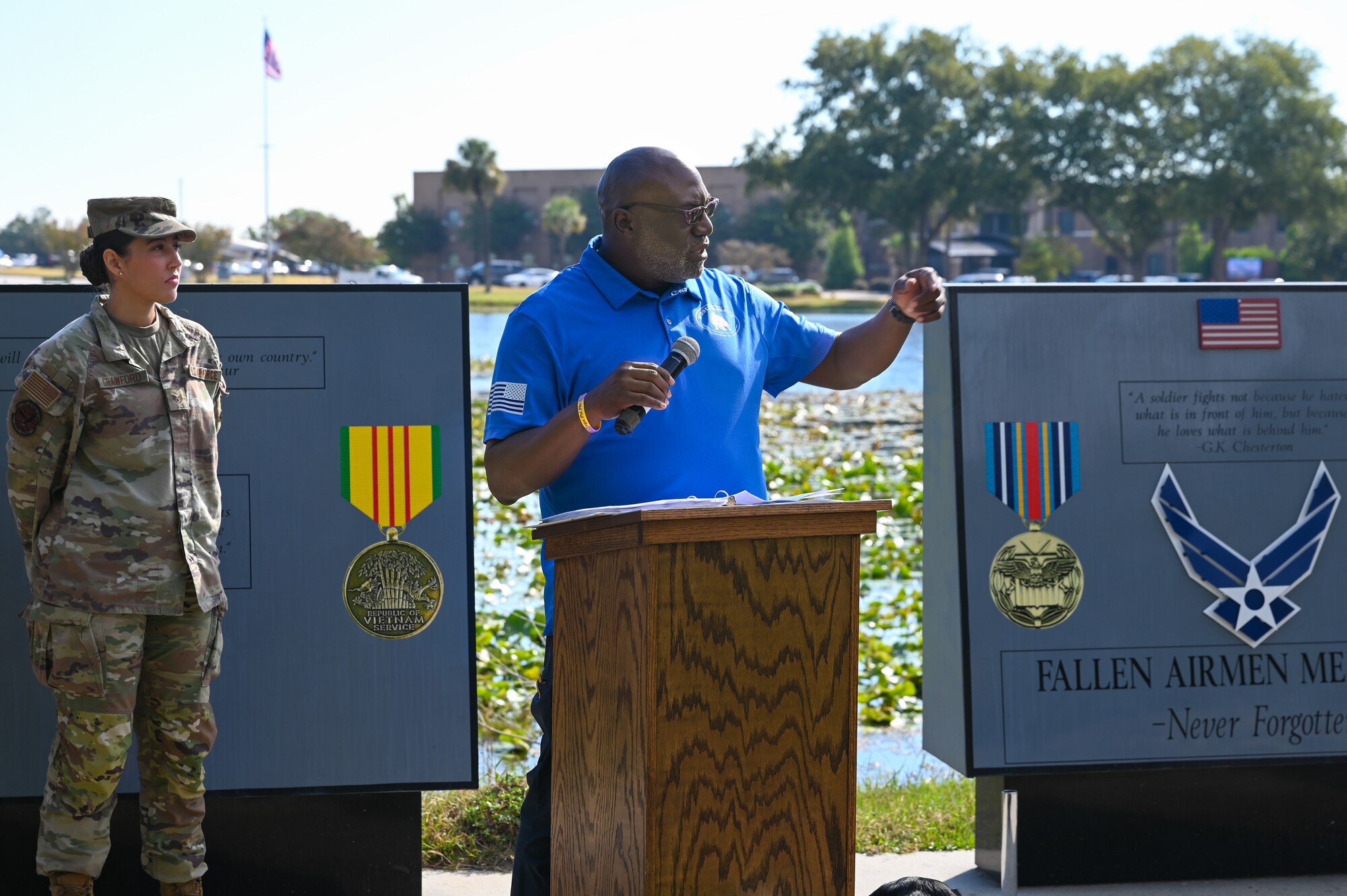 A man stands in front of a podium in the center of the photo, wearing sunglasses and a blue shirt, holding a microphone. His left arm is raised. Behind him are two memorial stones to either side and a female U.S. Air Force airman standing behind to the left.