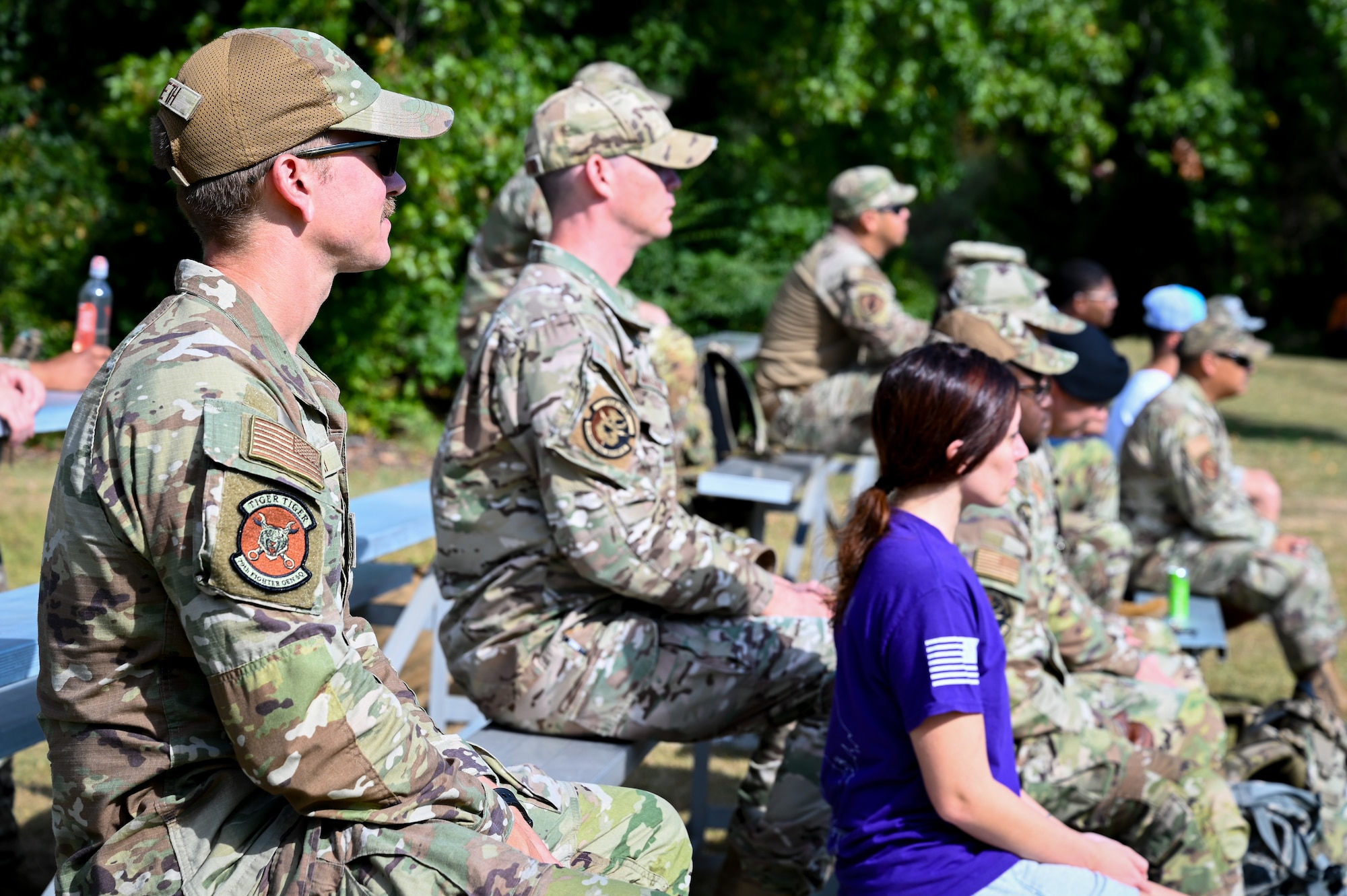 A group of airmen, one in civilian clothing, sit outside on bleachers facing the right.