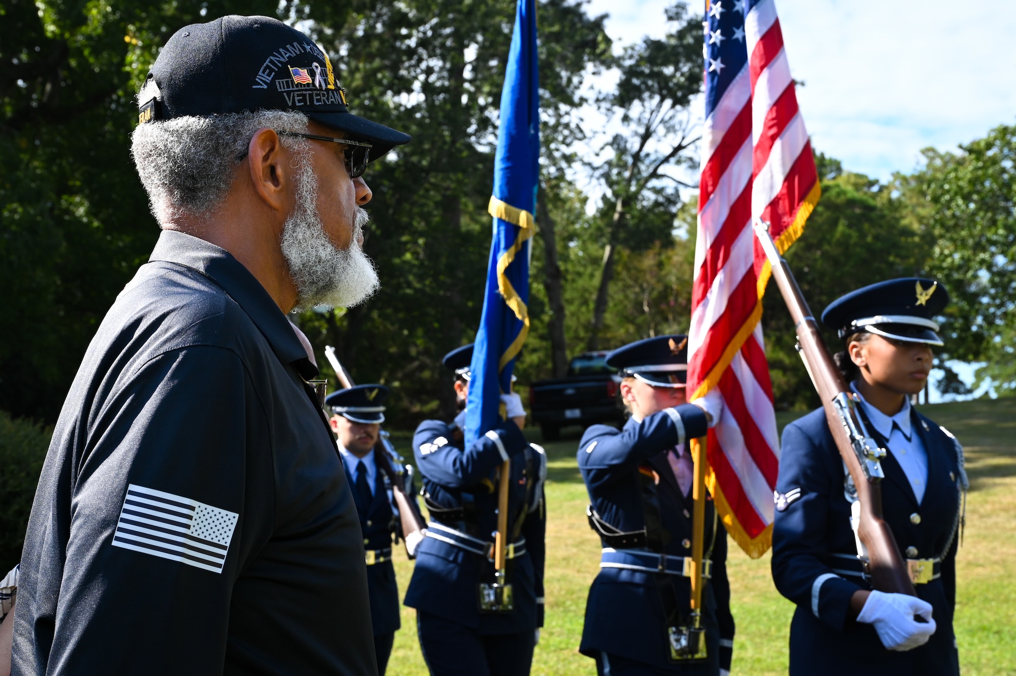 A man with a U.S. Air Force veteran hat stands to the left while members of a four-person U.S. Air Force honor guard detail march past.