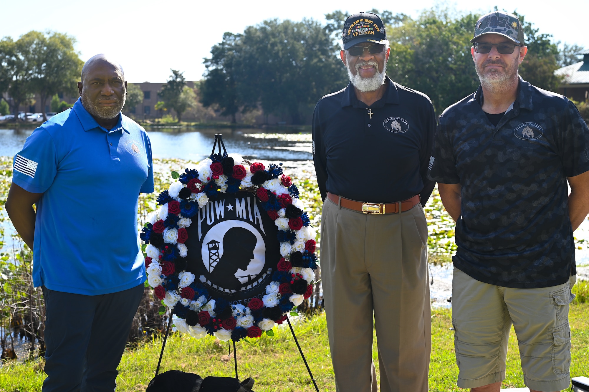 Three men gather around a POW/MIA commemoration plaque for a photo. One man has a veteran hat signifying that all three are veterans. They are outside with a pond in the background behind them. One is smiling, the other look serious.