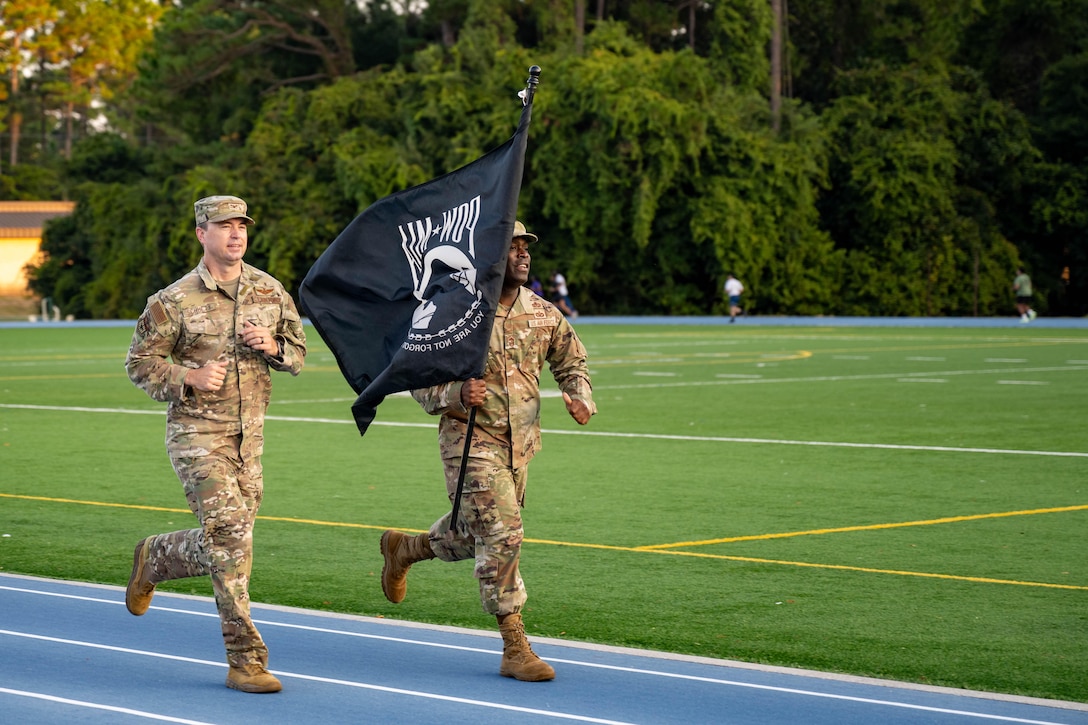 An airman holds a black flag with white writing and runs next to another airman on a blue track next a field with trees in the distance.