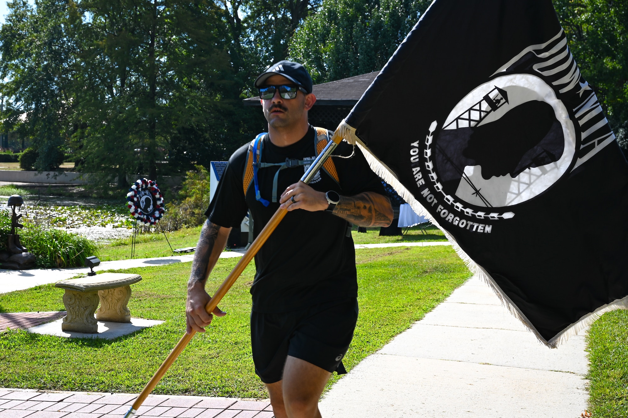 A man wearing a black shirt and shorts, hat, and sunglasses runs towards the camera carrying a POW/MIA guidon and flag. He is outside running along a sidewalk with a bench and the edge of a pond in the background.