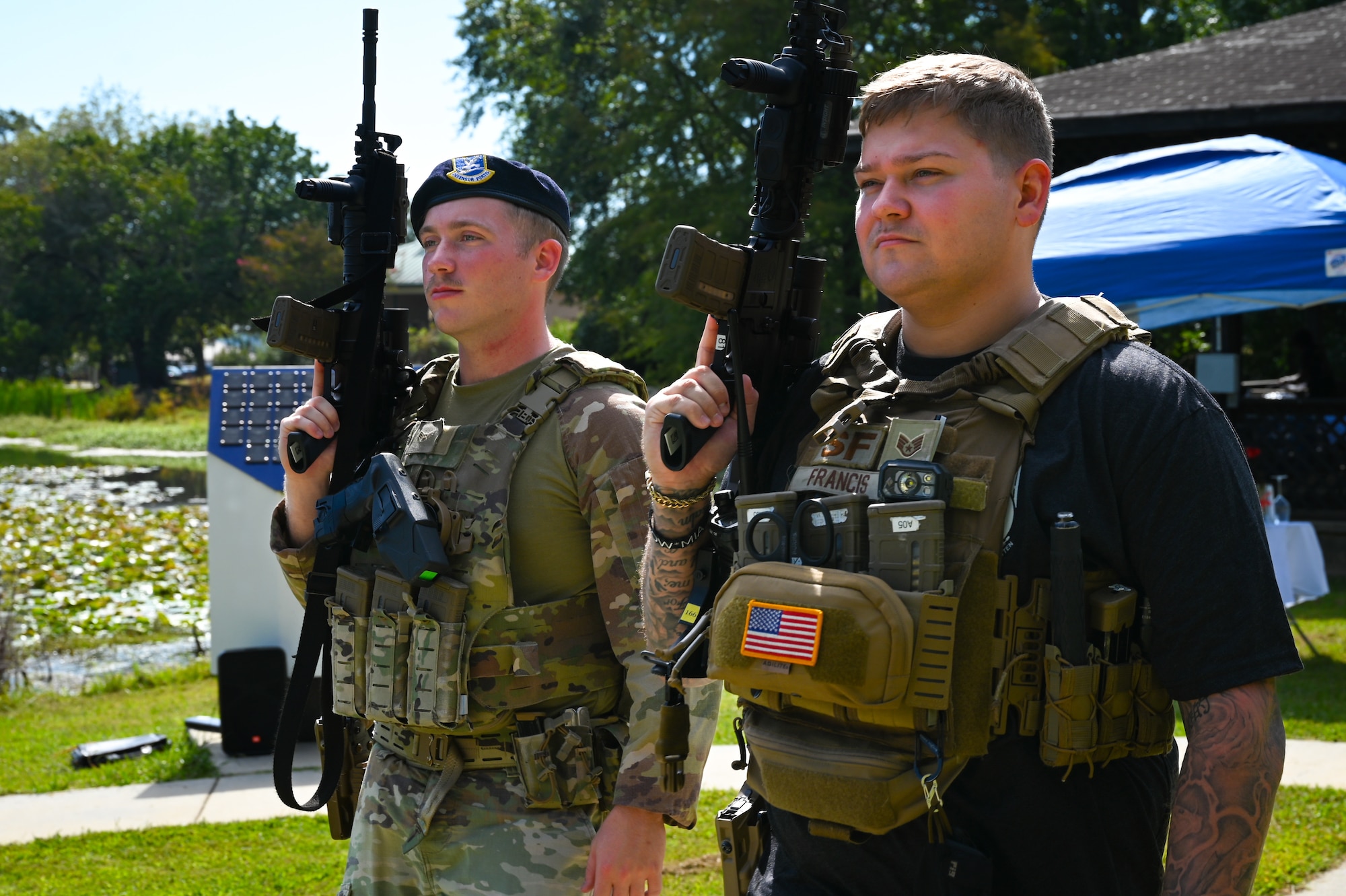 Two male Security Forces members, one in uniform and one in civilian clothing but both wearing Velcro battle vests, stand side by side holding training rifles, with the barrels pointing upward. There are trees, grass and the edge of a pond in the background.