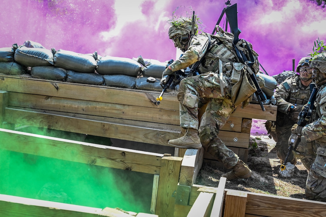 A guardsman in tactical gear and camouflage steps onto a wooden trench line as two others watch, surrounded by purple and green smoke.