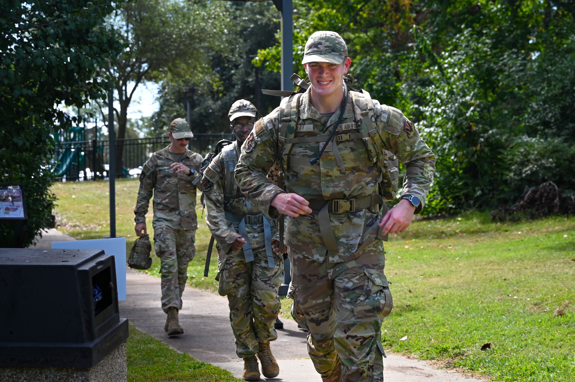Three US Air Force airmen approach in uniform in a line along a sidewalk as part of a 24-hour POW/MIA run/ruck vigiil. It is daytime and there is grass and trees in the background.