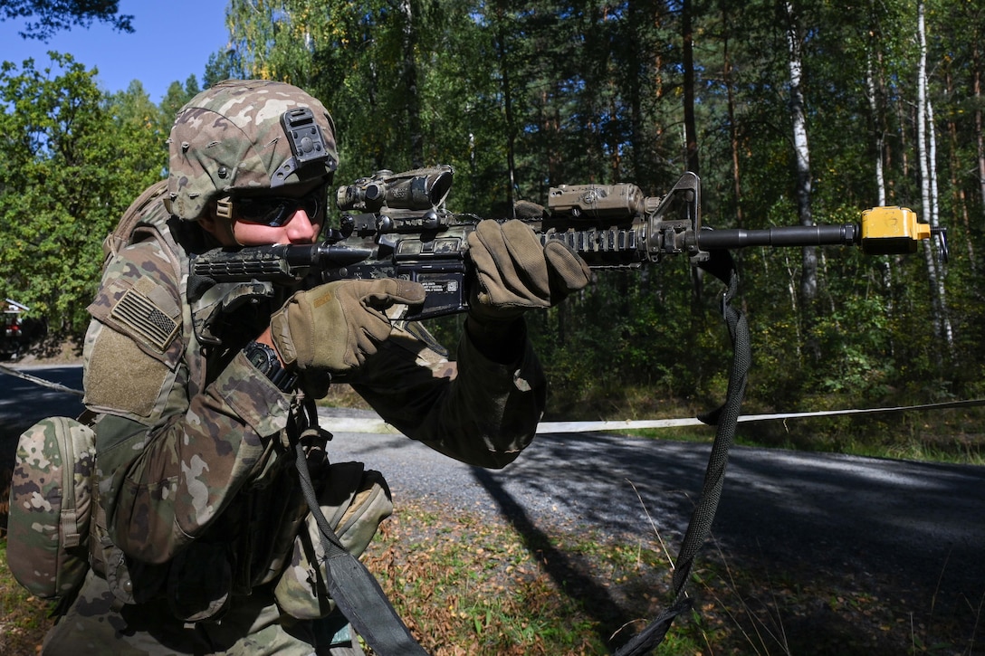 A soldier in tactical gear aims a service weapon to the right near an empty road running through a forest setting.