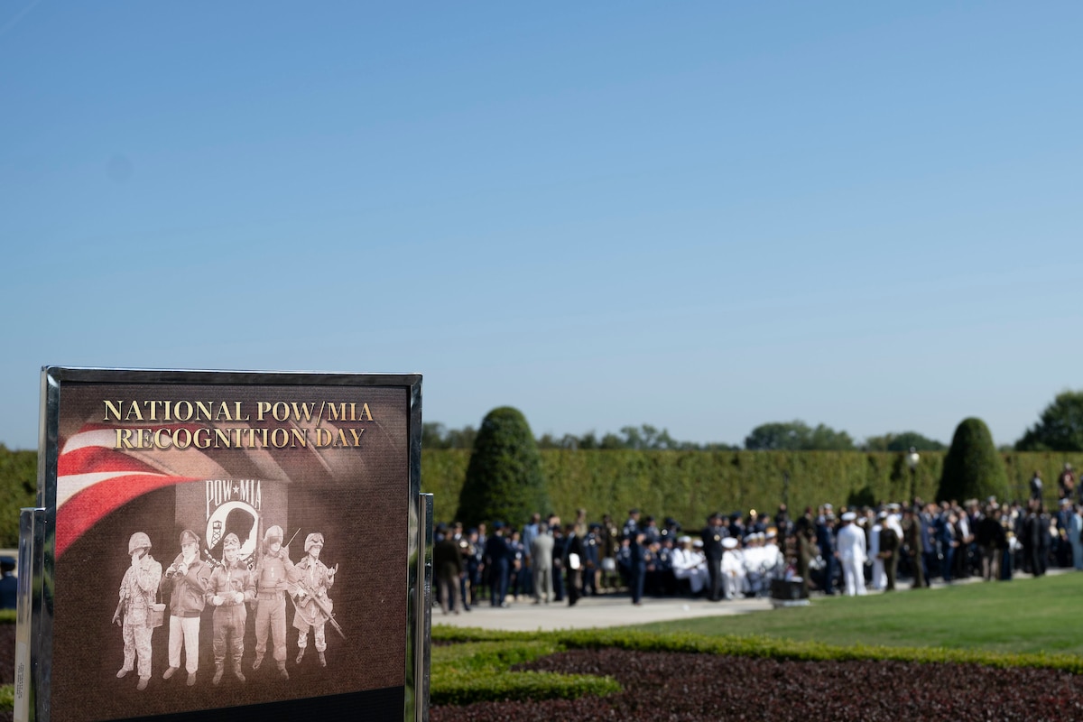 An outdoor graphic that shows service members from prior eras reads "National POW/MIA Recognition Day." A blurred crowd of people in formal civilian attire and dress military uniforms is in the background.