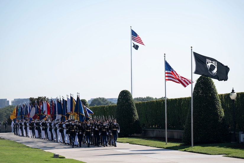 A large formation of service members in dress uniforms is marching along a cement walkway while holding up large flags from numerous U.S. states. They are walking alongside a large, green hedge and are passing by the American POW/MIA flags that are waving in the wind.