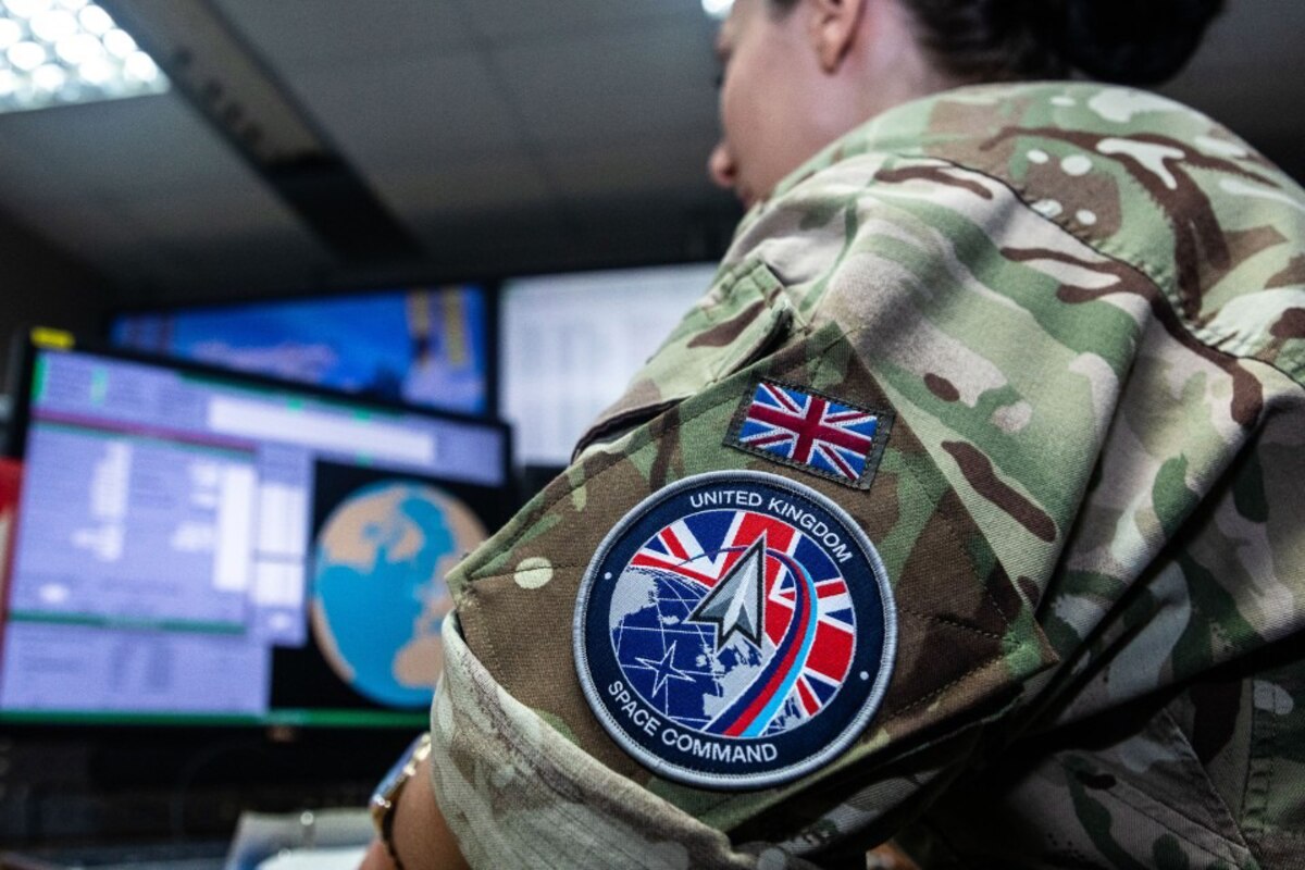 A person wearing a camouflage uniform with a U.K. Space Command patch sits at a computer.
