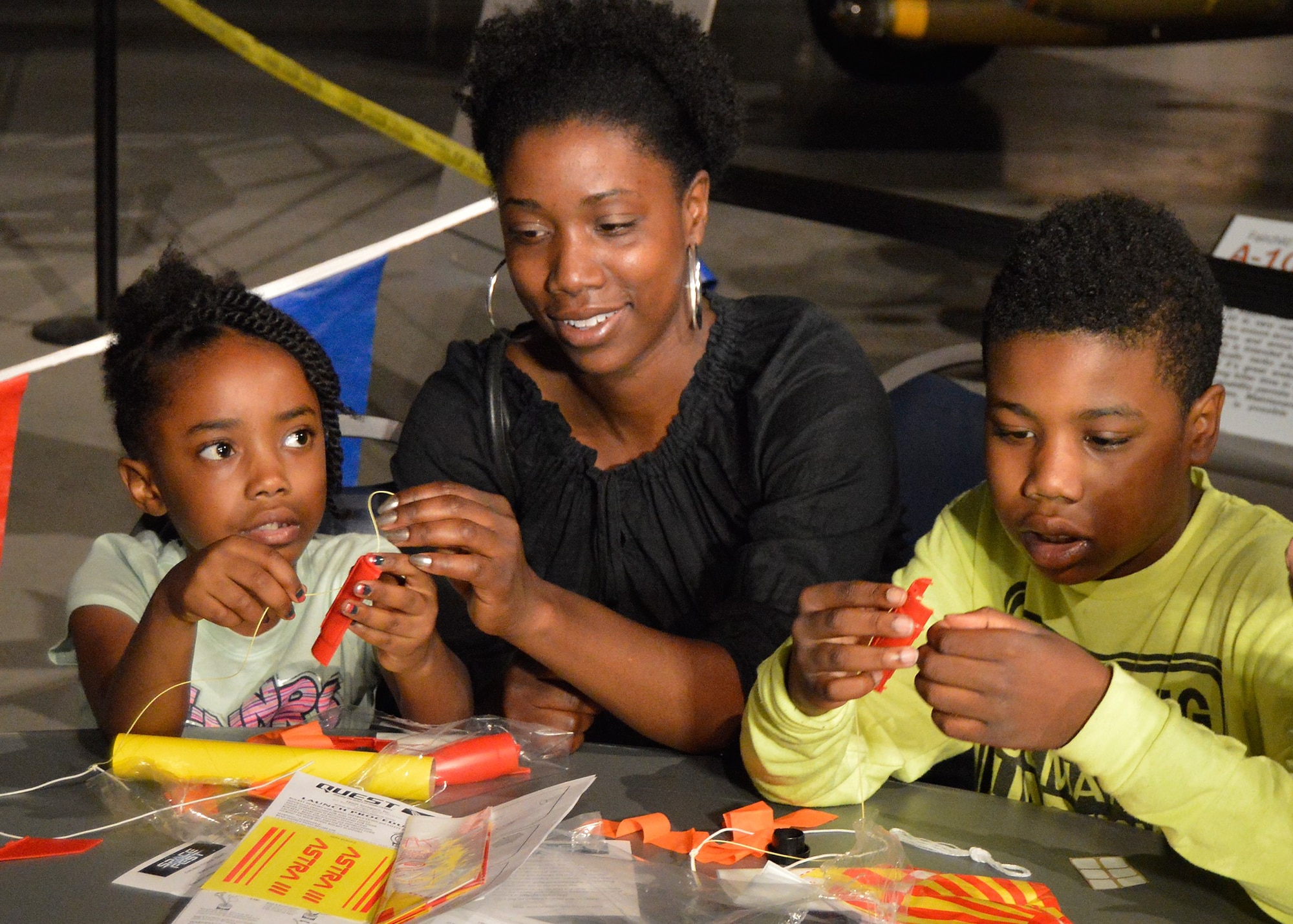 A woman and her two young children sitting at a table inside the National Museum of the Air Force building their rockets for launch on the front lawn of the museum.
