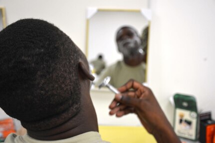 A person with shaving cream on their face looks into a mirror and shaves.