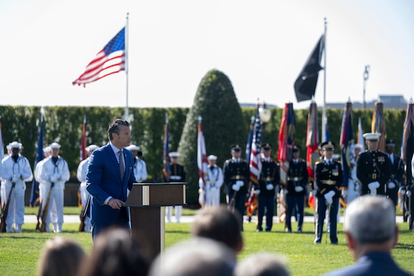 A man in civilian business attire is standing and speaking behind a lectern outdoors. A large group of military members in dress uniform attire are standing in the background, along with an American flag and a POW/MIA recognition flag.