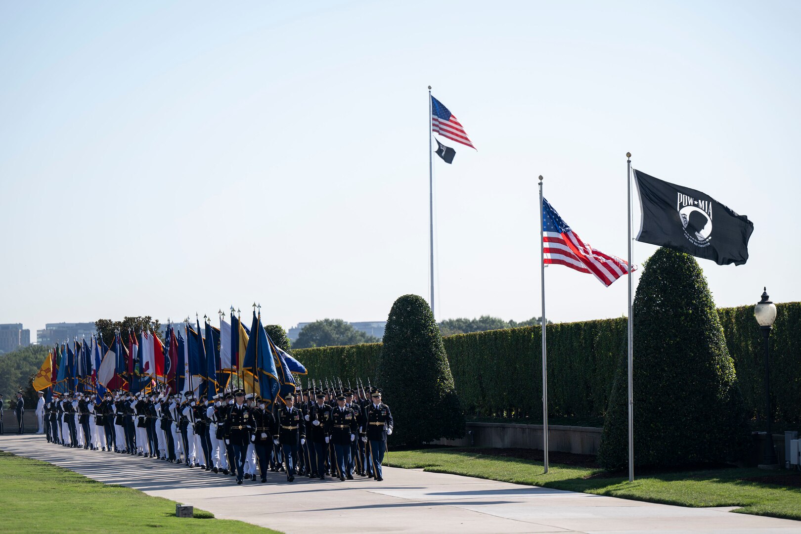 A large formation of service members in dress uniforms is marching along a cement walkway while holding up large flags from numerous U.S. states. They are walking alongside a large, green hedge and are passing by the American POW/MIA flags that are waving in the wind.
