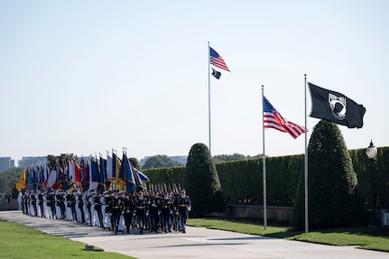 A large formation of service members in dress uniforms is marching along a cement walkway while holding up large flags from numerous U.S. states. They are walking alongside a large, green hedge and are passing by the American POW/MIA flags that are waving in the wind.