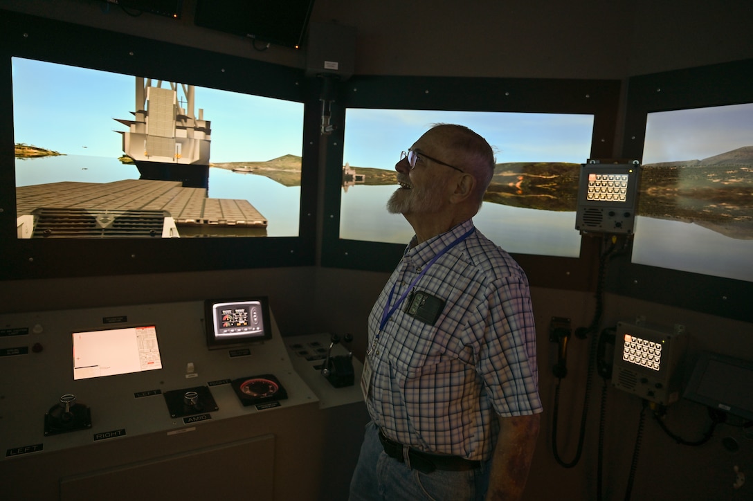 A man stands in a dark room looking at screens that simulate being on a boat and looking through the windows.