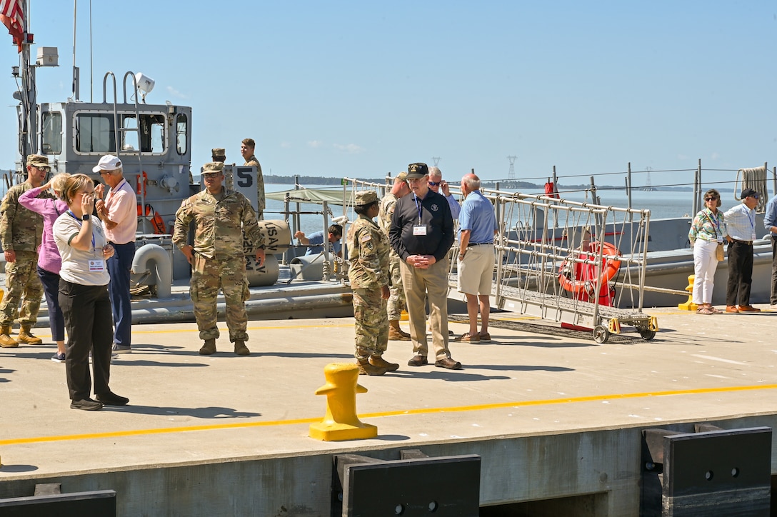 Civilians and Soliders stand on a dock by the water, talking in small groups. Its a sunny day.