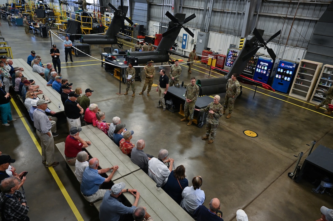 A short haired blonde woman stands before a room of Vietnam veterans. At her back are large portions of helicopters.