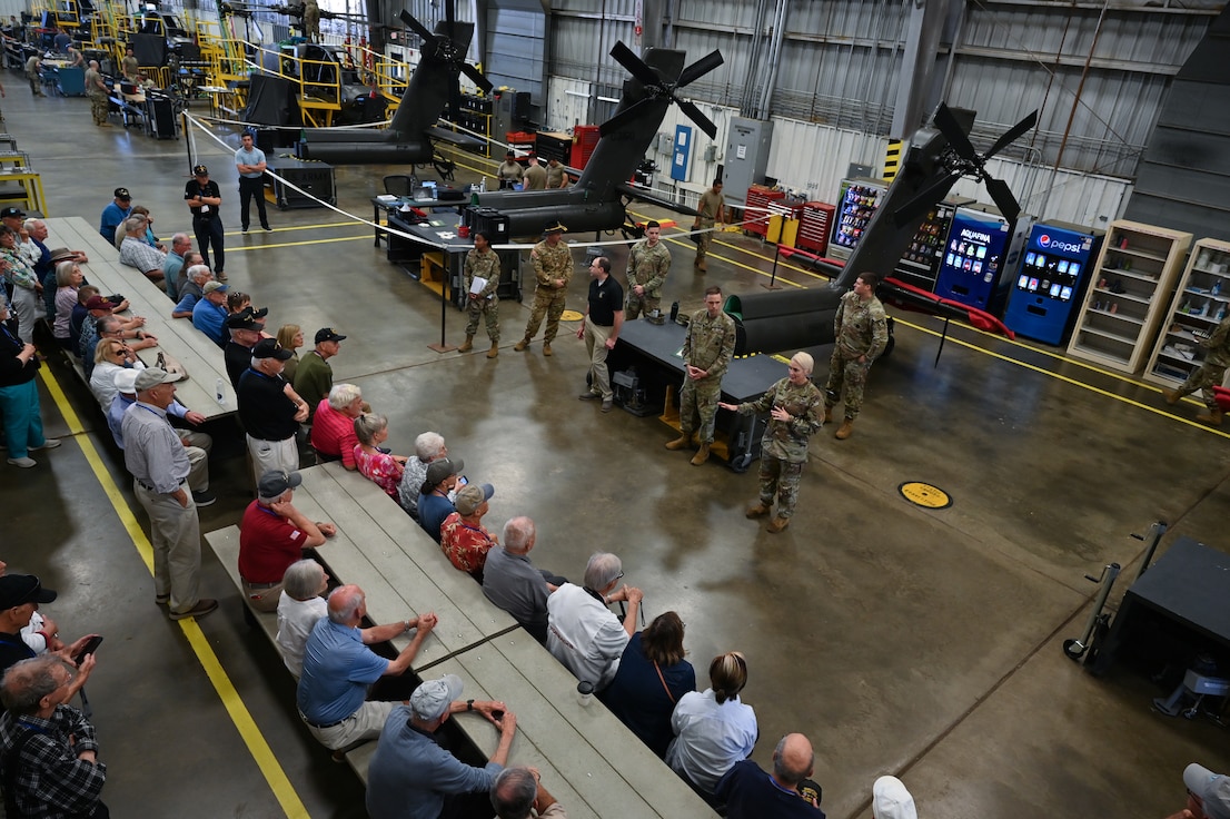 A short haired blonde woman stands before a room of Vietnam veterans. At her back are large portions of helicopters.