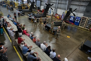 A short haired blonde woman stands before a room of Vietnam veterans. At her back are large portions of helicopters.