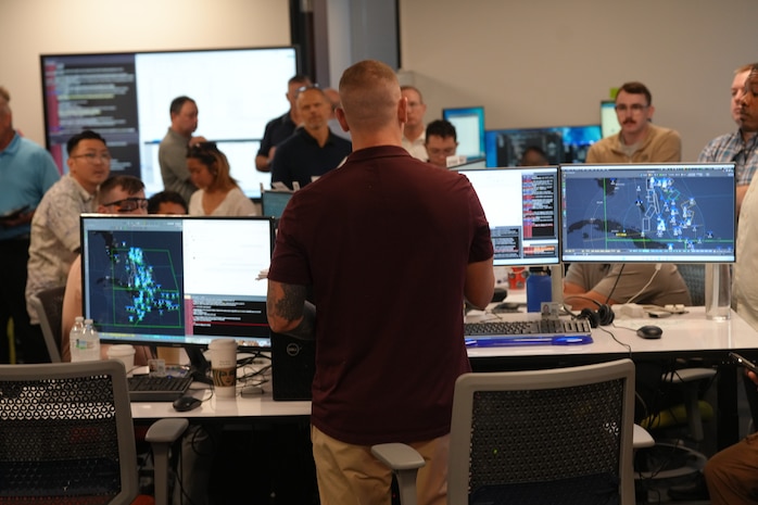 people sitting in chairs and standing listen to the leader standing, there is large computer screen mounted on the wall behind the people and people sitting working on computers in the room