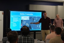 civilians sitting in chairs and standing listen to the leader standing, there is large computer screen mounted on the wall behind the people and people sitting working on computers in the room